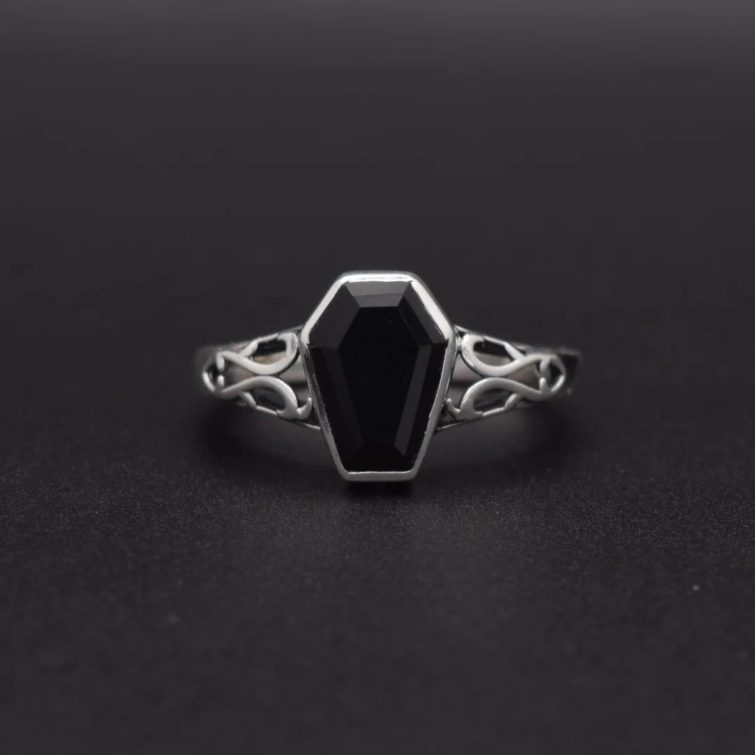 A gothic coffin-shaped ring with a faceted black gemstone and ornate silver filigree band, set against a dark gray background.