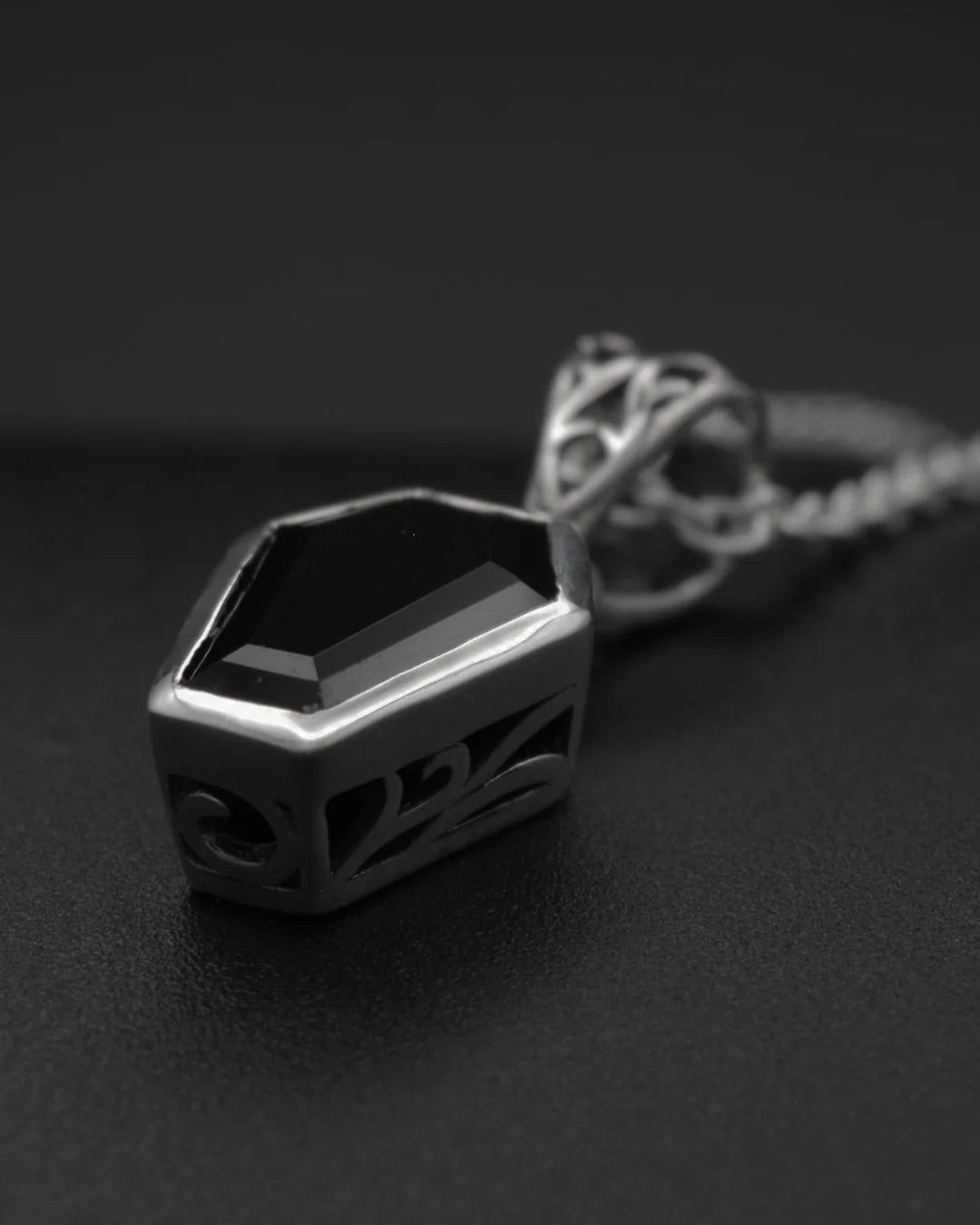 A gothic-style silver pendant necklace featuring a faceted black gemstone in a geometric, coffin-like frame with intricate cutout details, resting on a dark textured surface.
