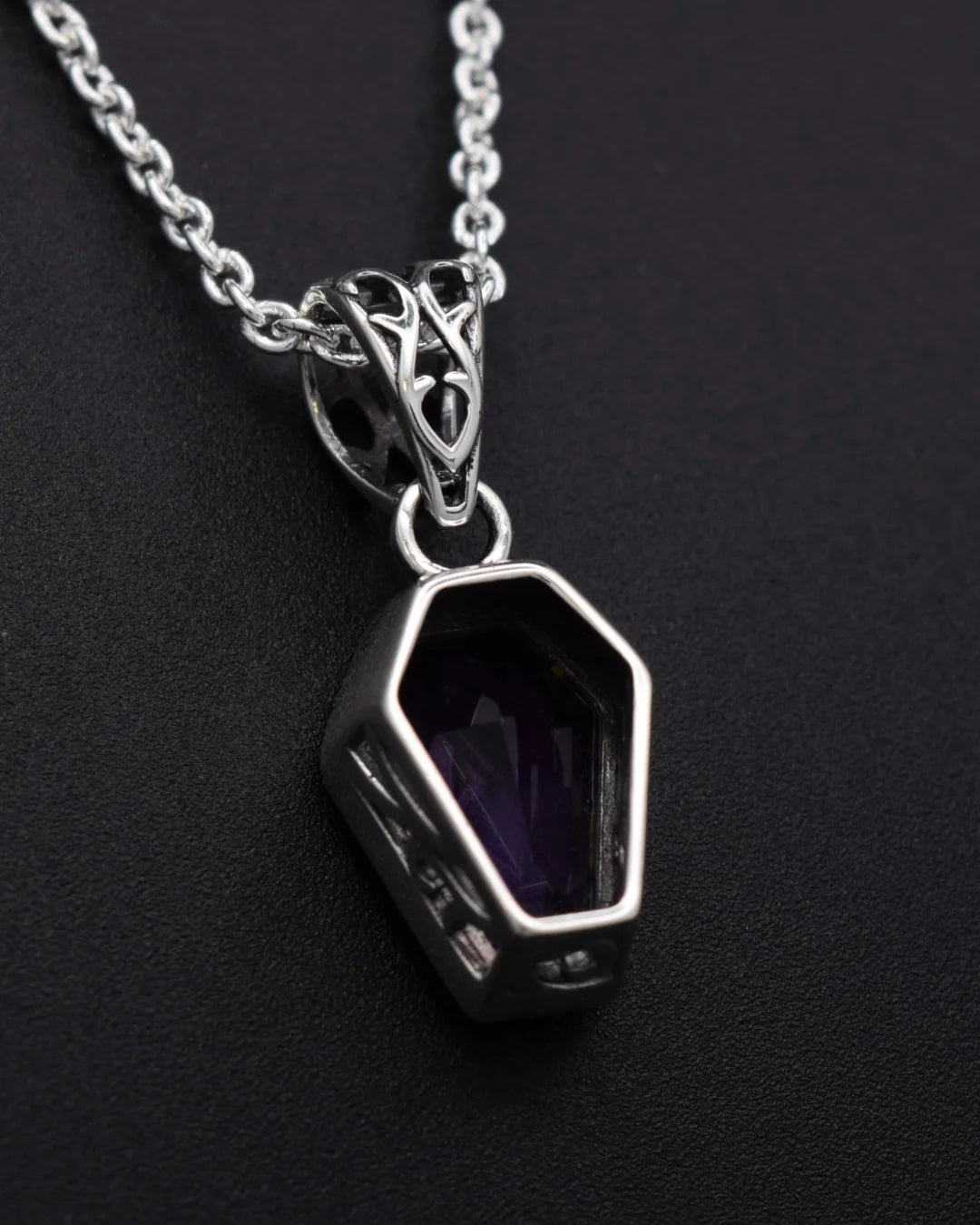 A silver coffin-shaped pendant necklace with a faceted purple gemstone, shown on a dark textured background.