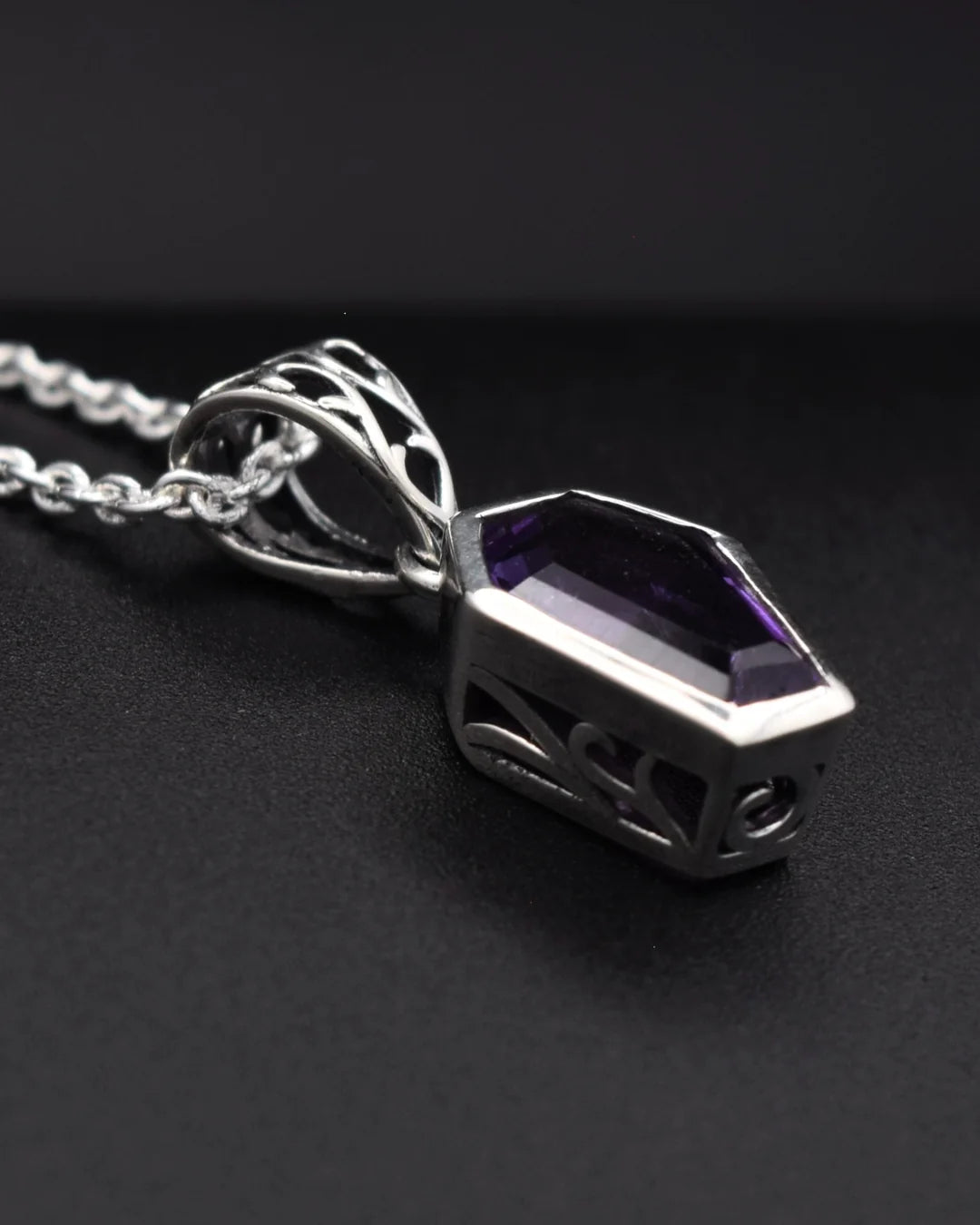 A silver pendant necklace featuring a faceted purple gemstone in a geometric, openwork metal setting, suspended from a silver chain against a dark background.