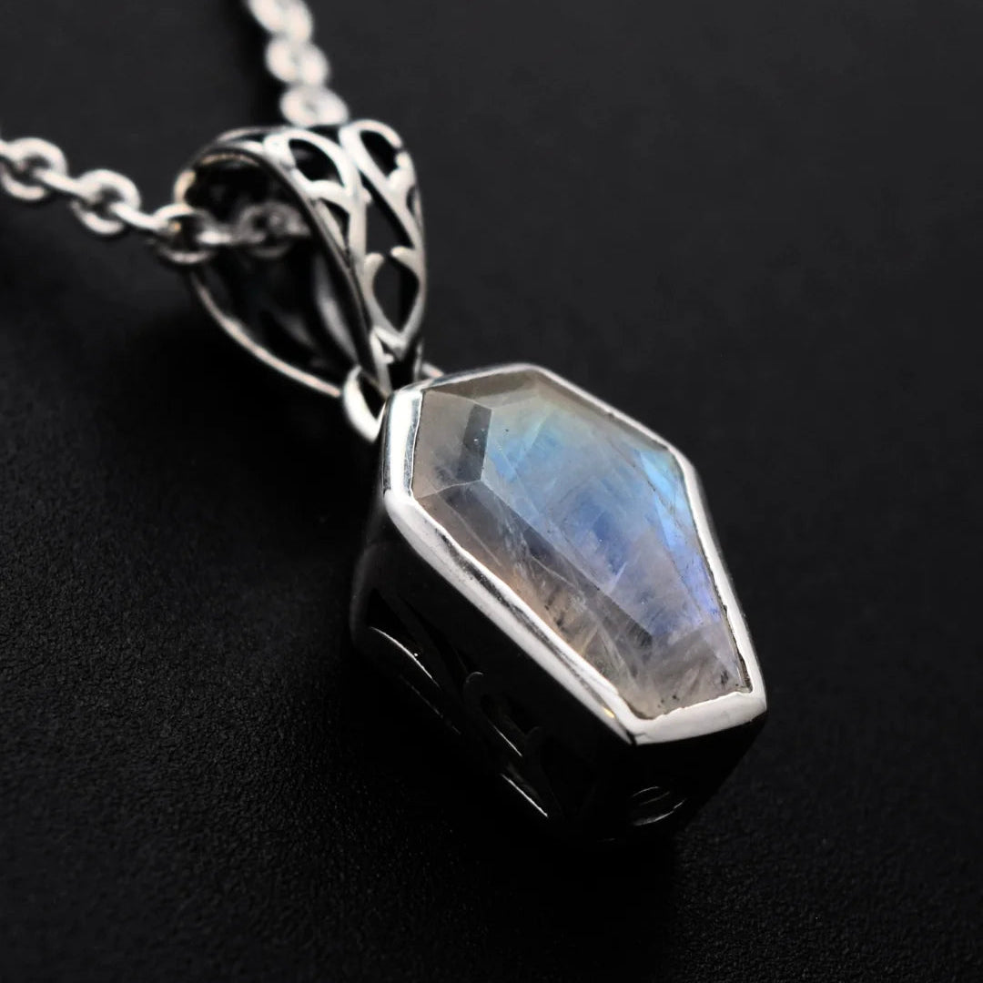 A silver pendant necklace featuring a faceted, coffin-shaped iridescent gemstone with blue and green flashes, set in a polished silver bezel with an ornate filigree bail, resting on a textured black surface.
