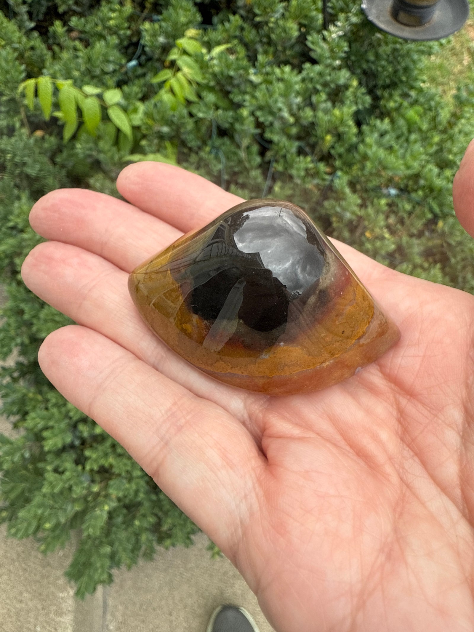 Hand holding a polished crystal stone with a dark center and yellowish-brown exterior, outdoors.