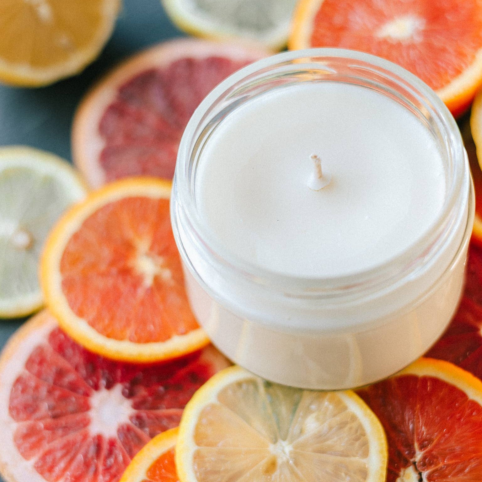 White candle in a glass jar surrounded by sliced citrus fruits on a dark surface