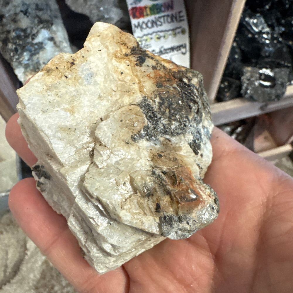 Hand holding a rough mineral specimen with a box of similar specimens in the background.