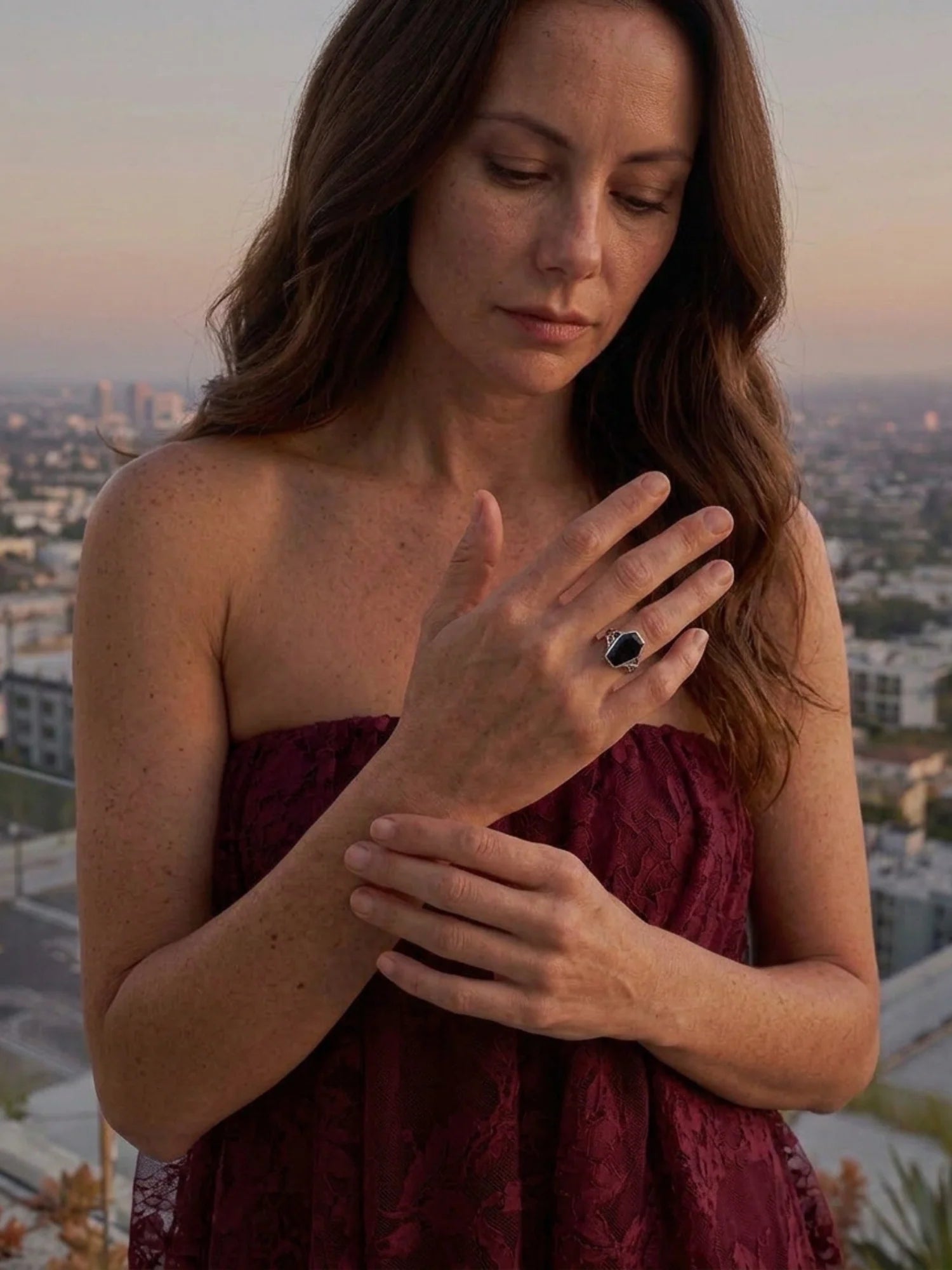 A woman wearing a strapless burgundy lace dress looks down at a ring she is wearing on her finger, with a blurred city skyline in the background at sunset.
