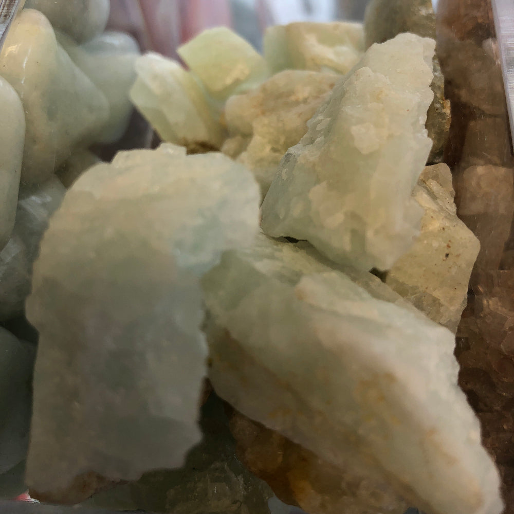 A collection of raw Aquamarine gemstones displayed in a bowl, with a white crystal behind them.