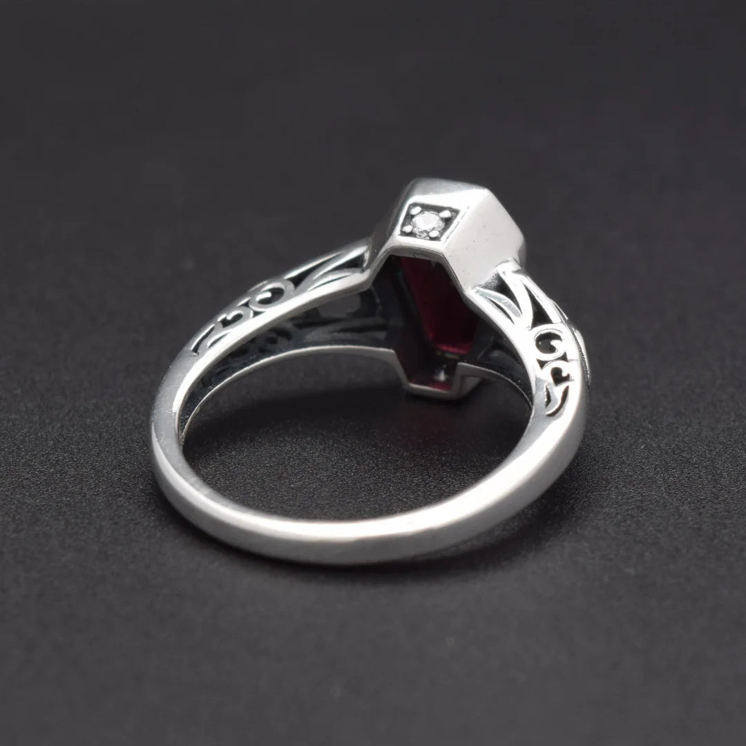 A silver ring featuring a geometric, faceted red gemstone centerpiece and a small clear stone on the upper band, set against a dark textured background.