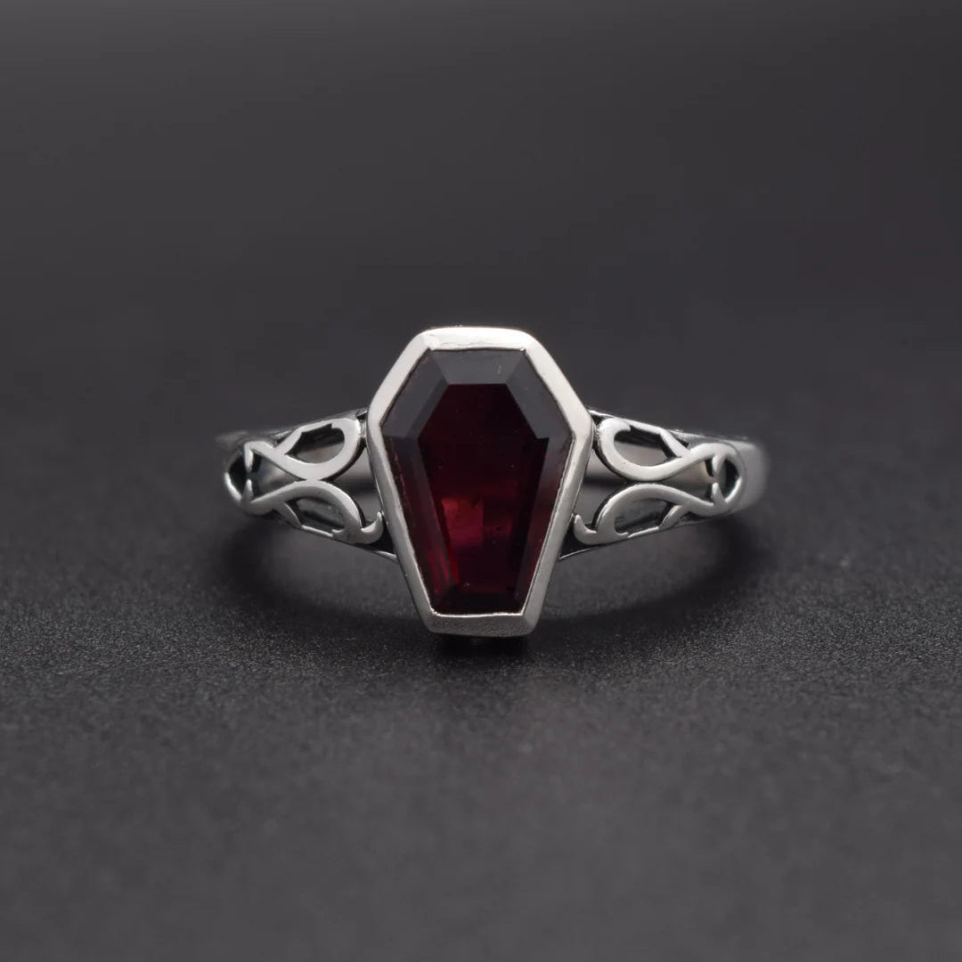 A gothic-style coffin-shaped ring featuring a deep red gemstone in a silver bezel setting, with an ornate Celtic knot band, shown on a dark gray background.