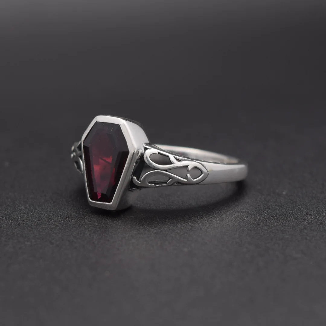 A silver gothic-style ring featuring a faceted dark red gemstone in a coffin-shaped bezel, set on a dark gray background.