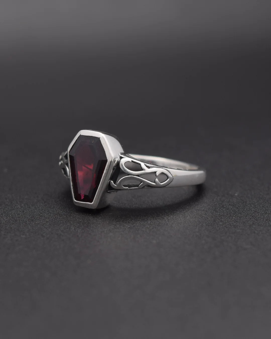 A silver gothic-style ring featuring a faceted dark red gemstone in a coffin-shaped bezel, set on a dark gray background.
