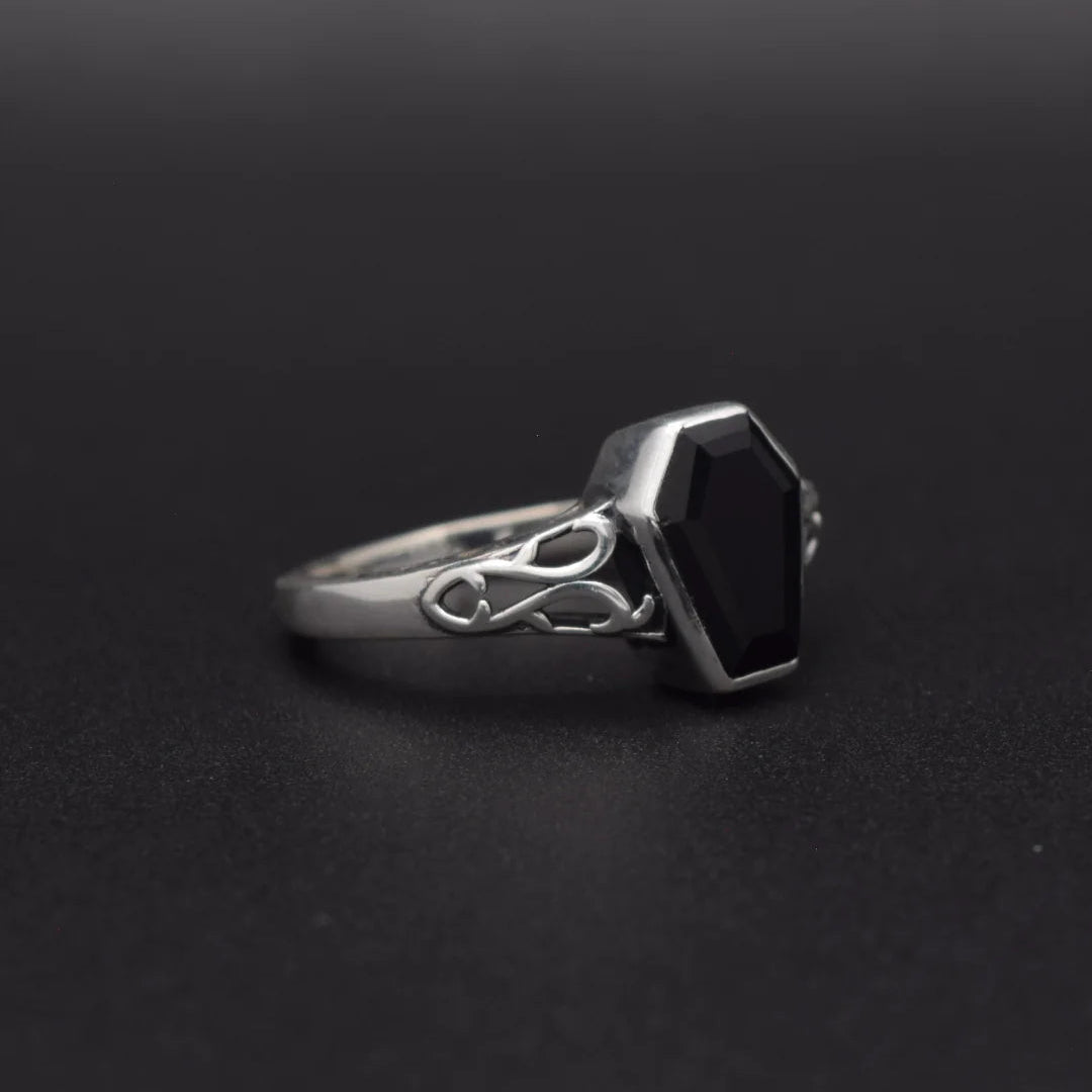A silver ring with a coffin-shaped black gemstone and an intricate Celtic knot design on the band, set against a dark gray background.