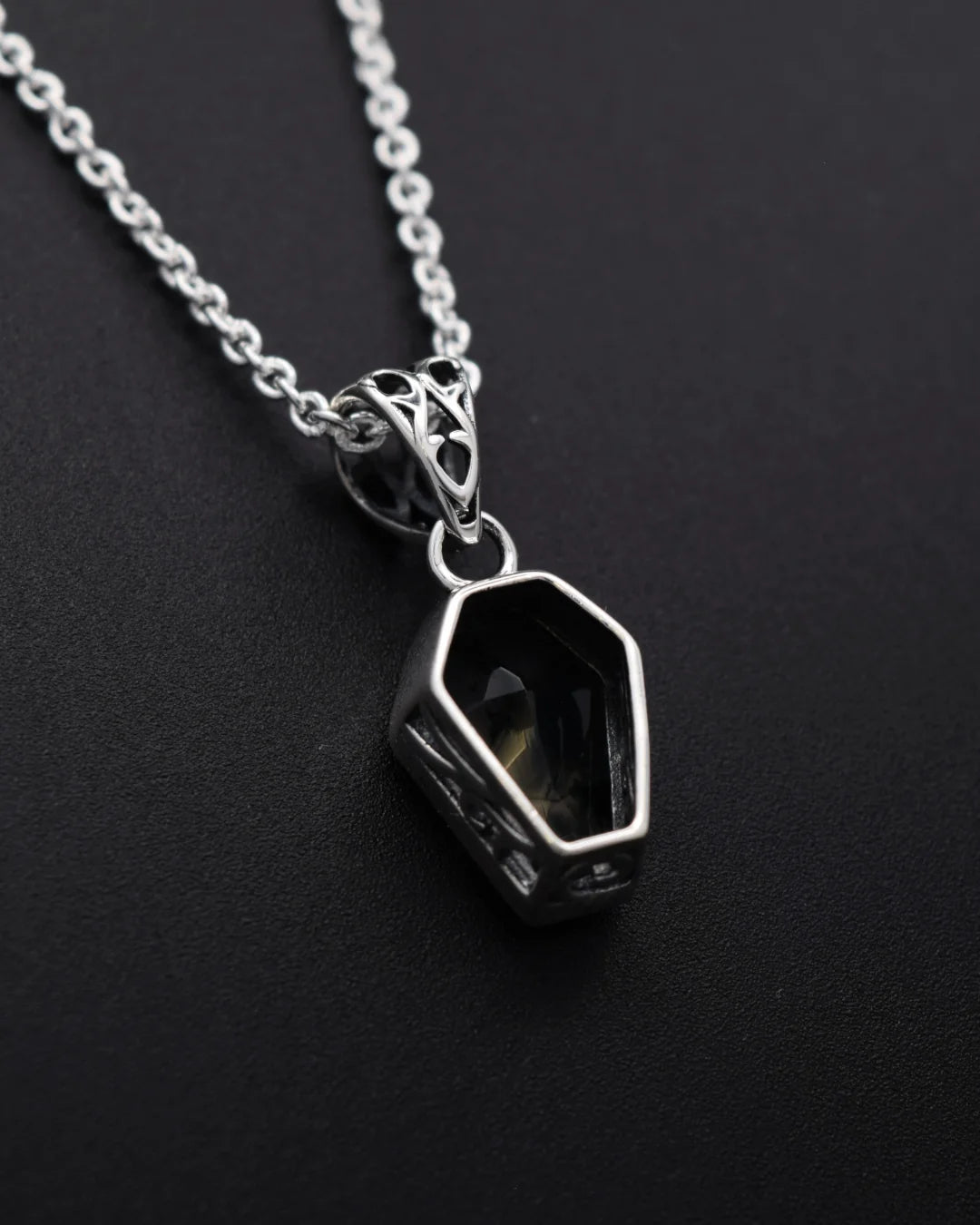 A silver-toned chain necklace with a geometric, coffin-shaped pendant featuring a dark, smoky center, displayed on a dark textured background.