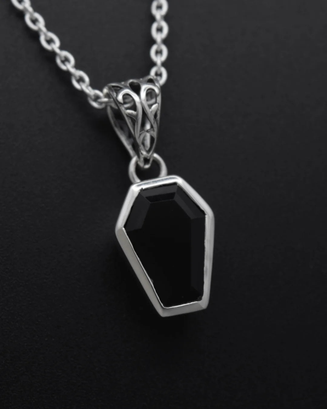 A silver coffin-shaped pendant necklace with a faceted black gemstone, shown on a dark textured background.