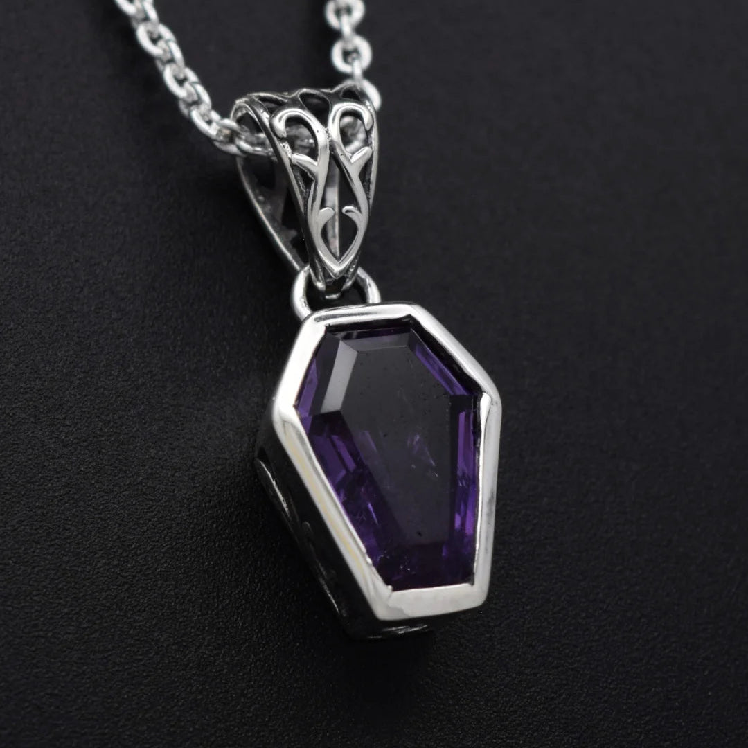 A silver pendant necklace featuring a faceted coffin-shaped purple gemstone, displayed on a textured black background.
