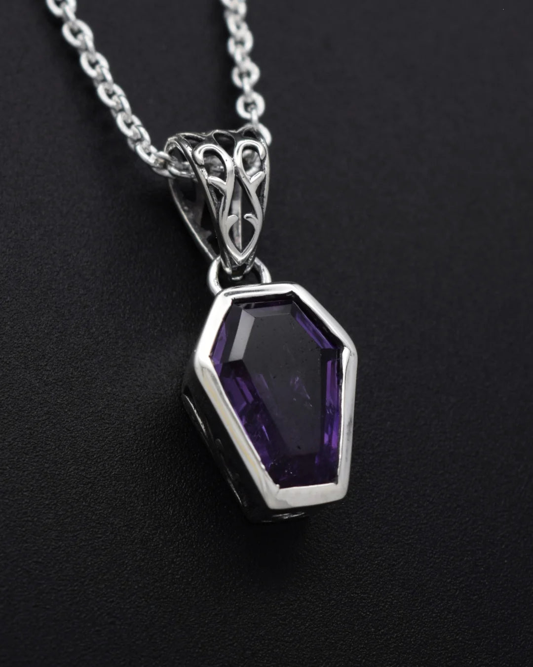 A silver pendant necklace featuring a faceted coffin-shaped purple gemstone, displayed on a textured black background.