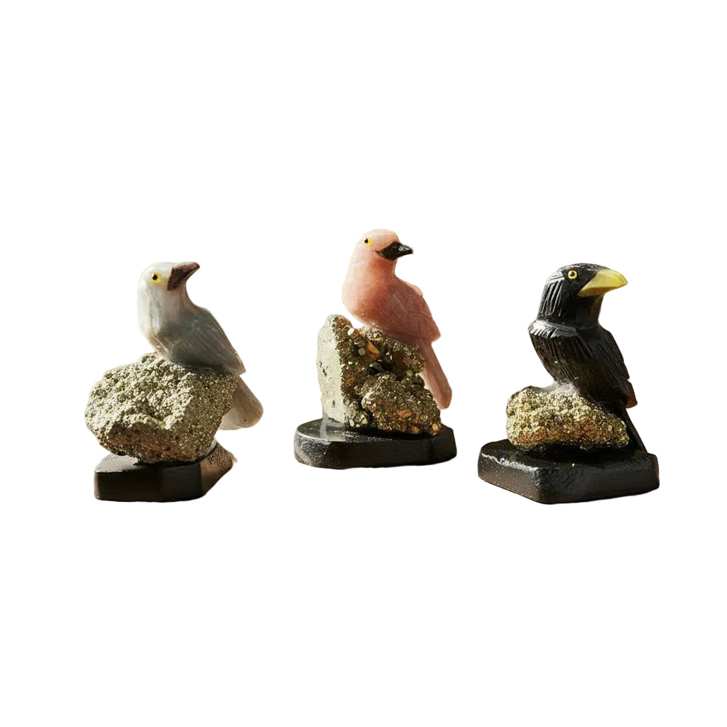 Three small bird figurines perched on metallic rock-like bases, displayed on a plain white background. The set includes a pink bird, a gray bird, and a black bird, each with yellow eyes and mounted on black square bases.