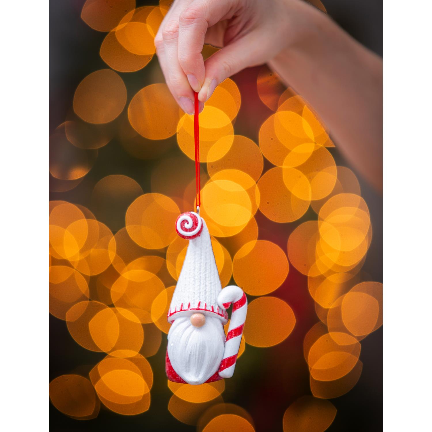 Hand holding a bearded candy cane gnome ornament by its red ribbon in front of glowing Christmas lights