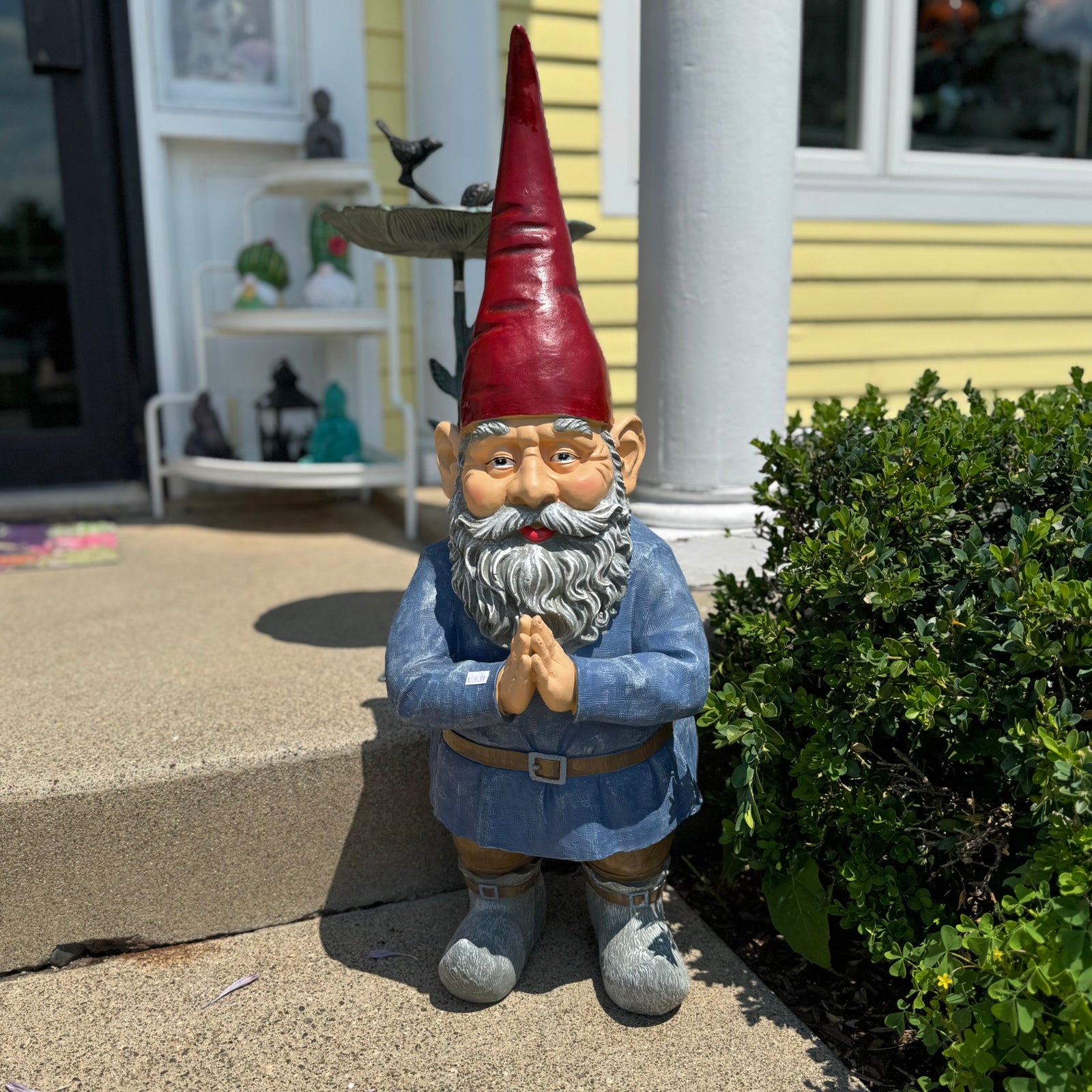 A painted garden gnome statue stands on a concrete porch step outdoors. The gnome has a tall red hat, gray beard, blue outfit, and gray shoes, with a small bird perched on the hat.