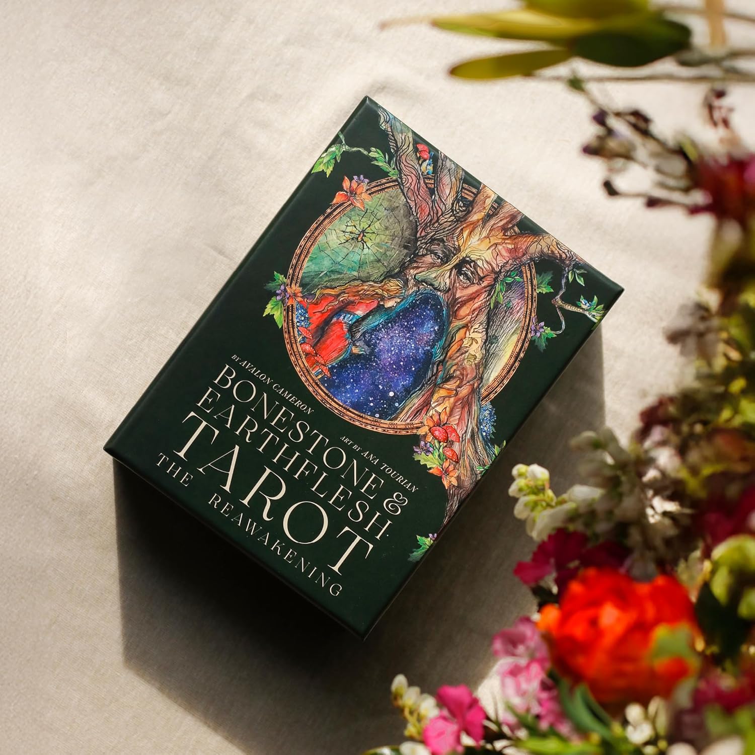 Bonestone & Earthflesh Tarot box with tree spirit artwork lying on linen beside fresh flowers