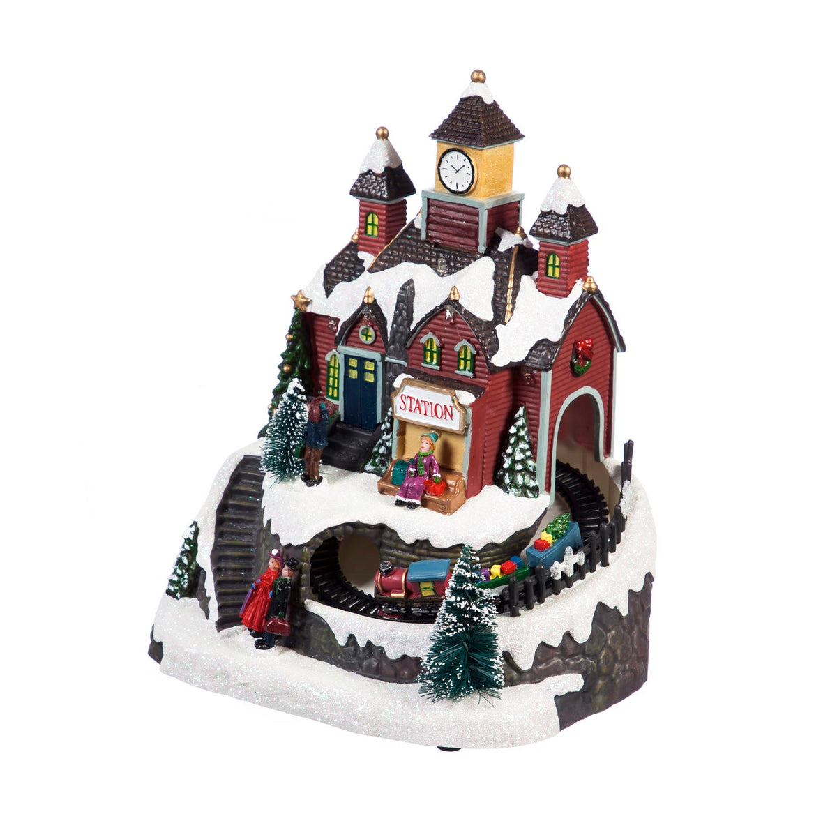 Light-up Christmas village train station with clock tower, snowy roofs, bottlebrush trees and a small train on a track below