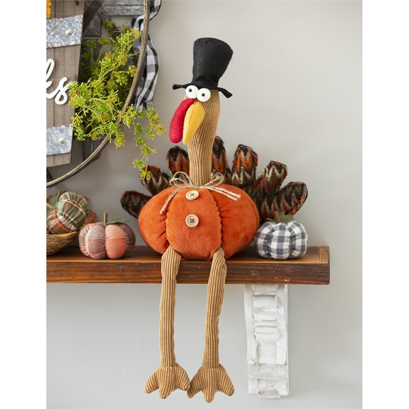 Gobbles the plush turkey sitting on a wooden fall mantel shelf with his long corduroy legs dangling over the edge, next to fabric pumpkins