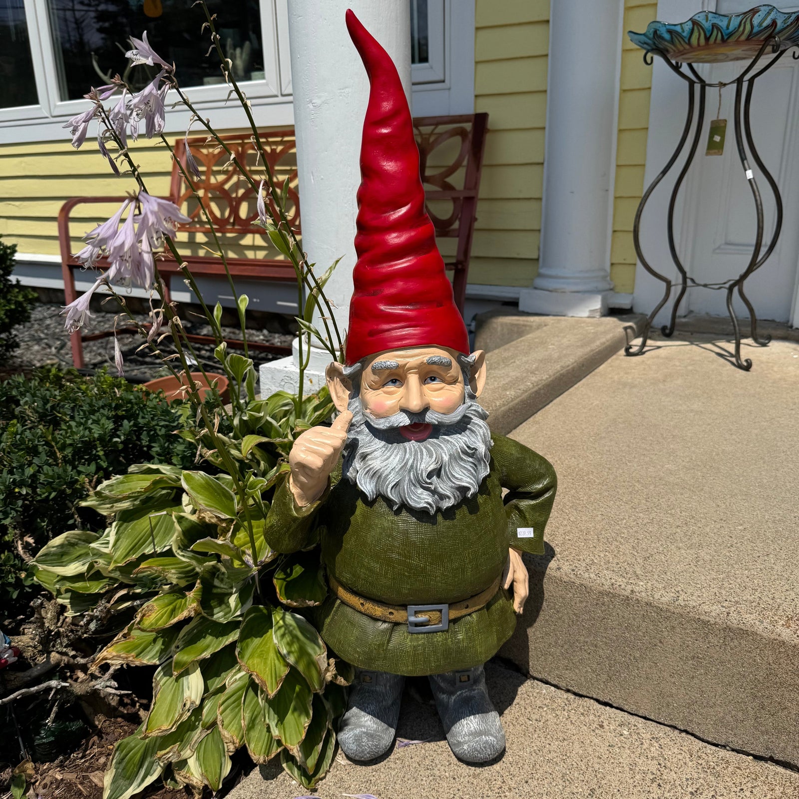 A decorative garden gnome statue standing outdoors on a concrete porch step beside green plants. The gnome has a tall red pointed hat, gray beard, olive green outfit, and gray shoes.