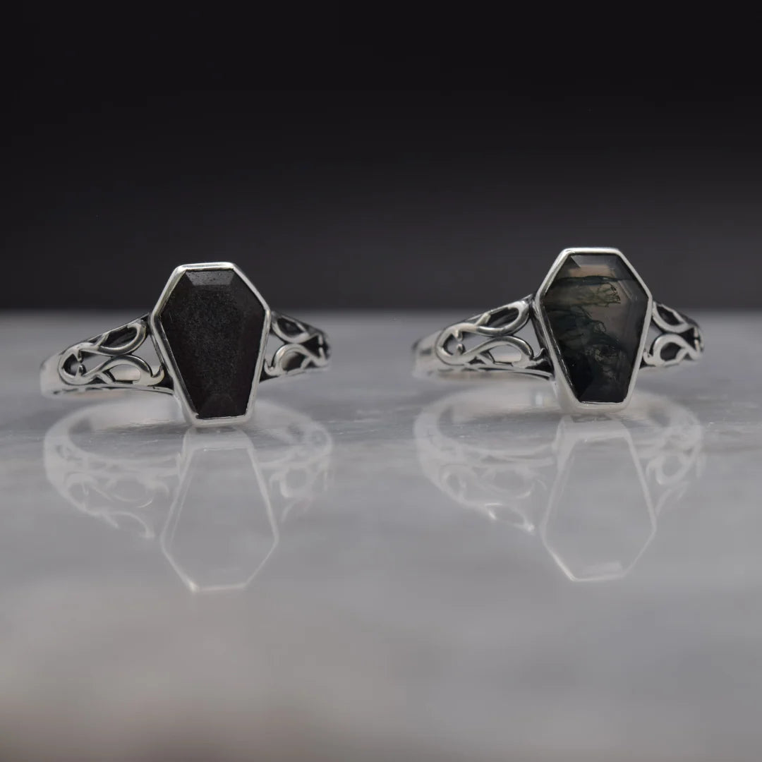 A pair of silver rings with hexagonal black stone inlays, displayed on a reflective gray surface against a dark background.