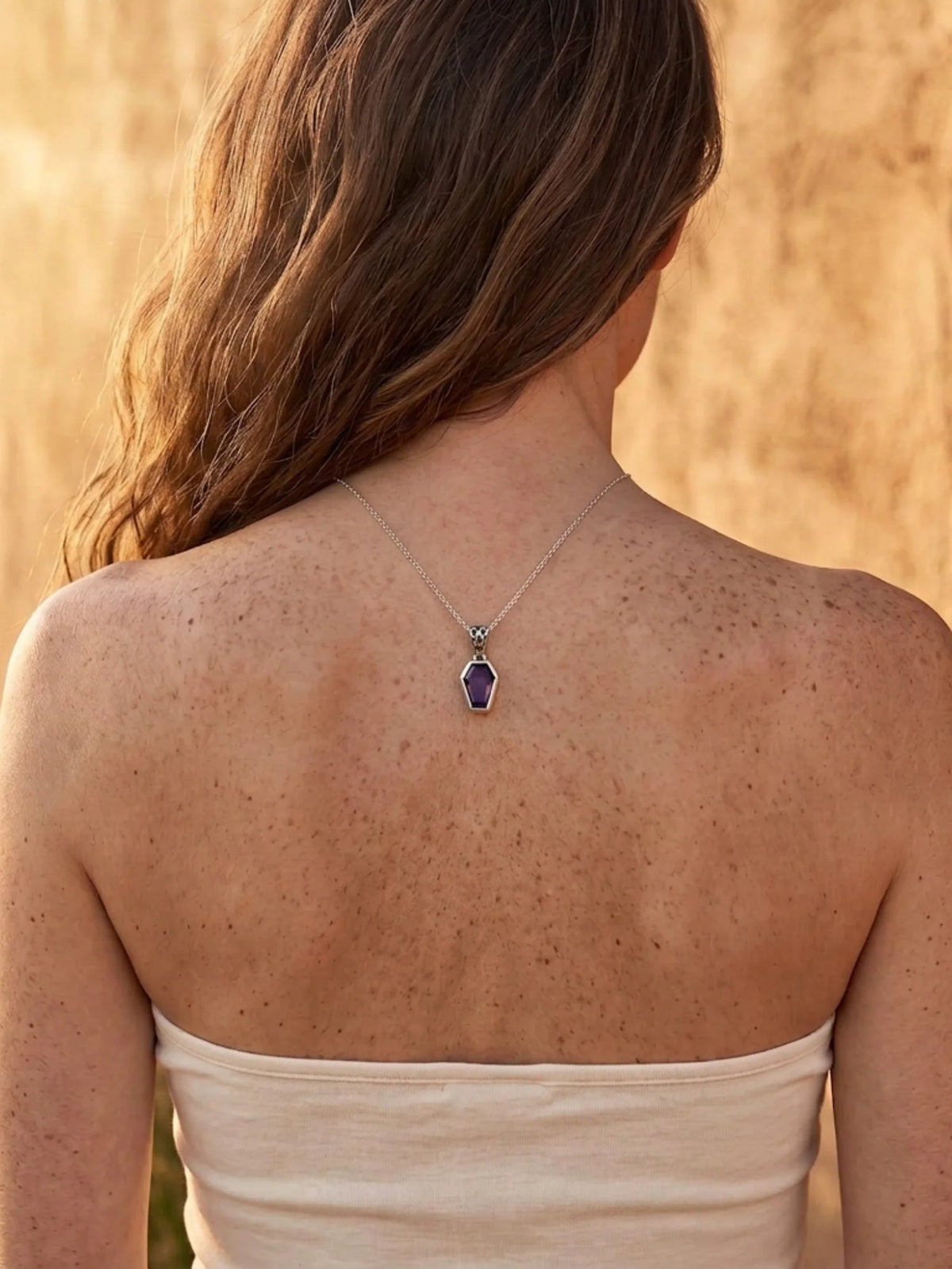 A woman shown from behind wearing a delicate silver chain necklace with a small teardrop-shaped dark purple pendant, set against a warm, textured beige background.