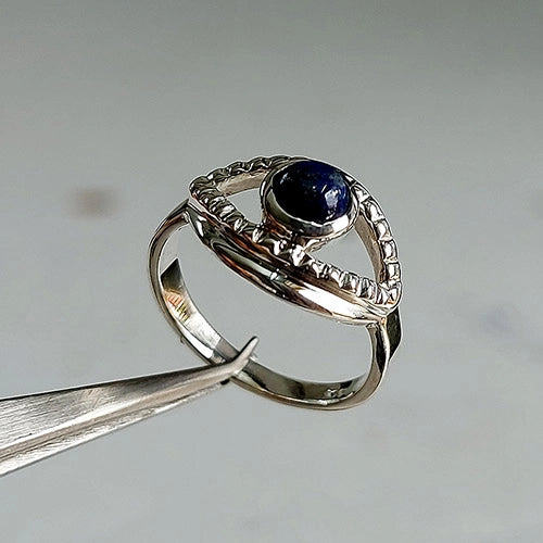 A silver ring with a dark blue oval gemstone, held by metal tweezers against a neutral light gray background.