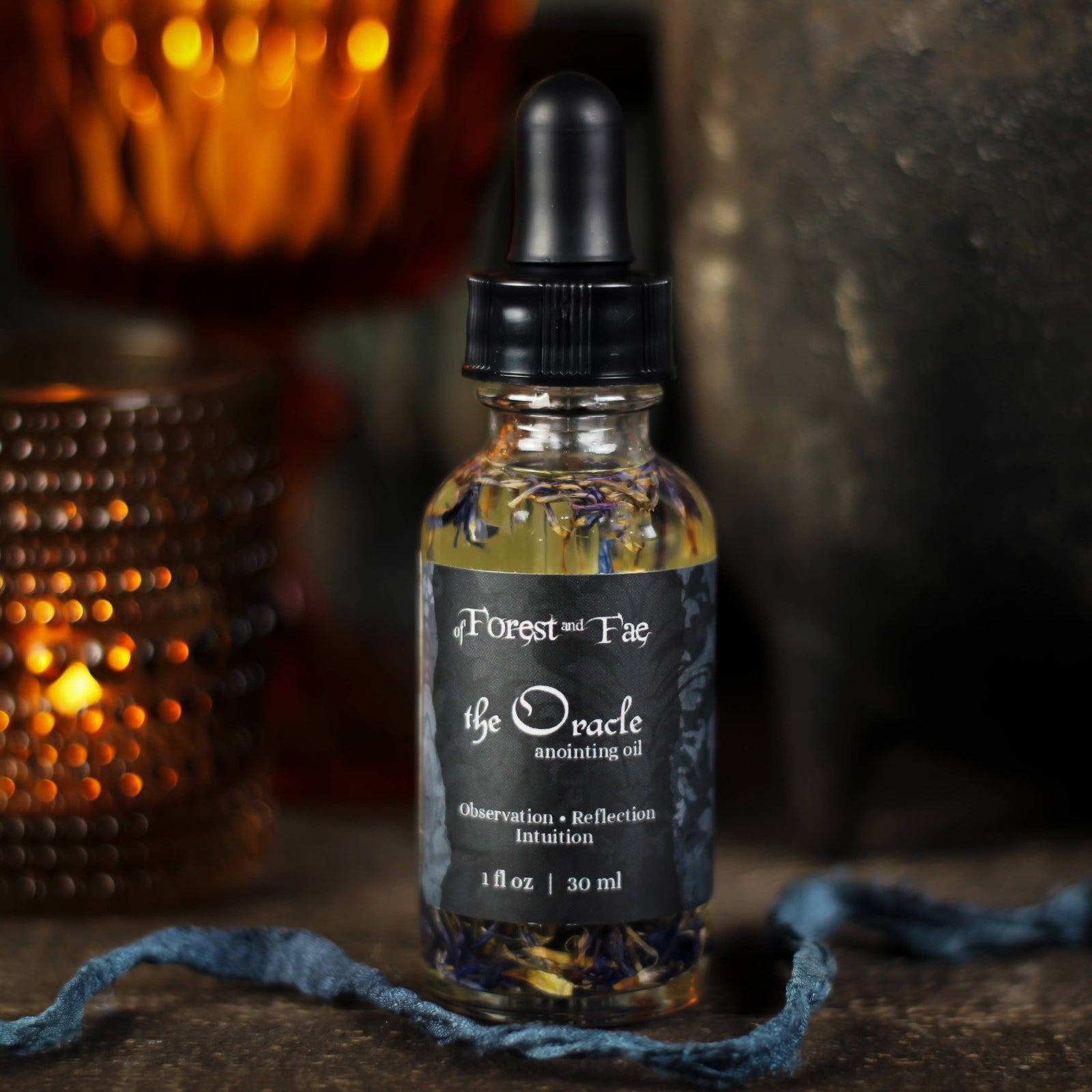 The Oracle anointing oil 1 fl oz dropper bottle with blue petals inside, beside a glowing candle