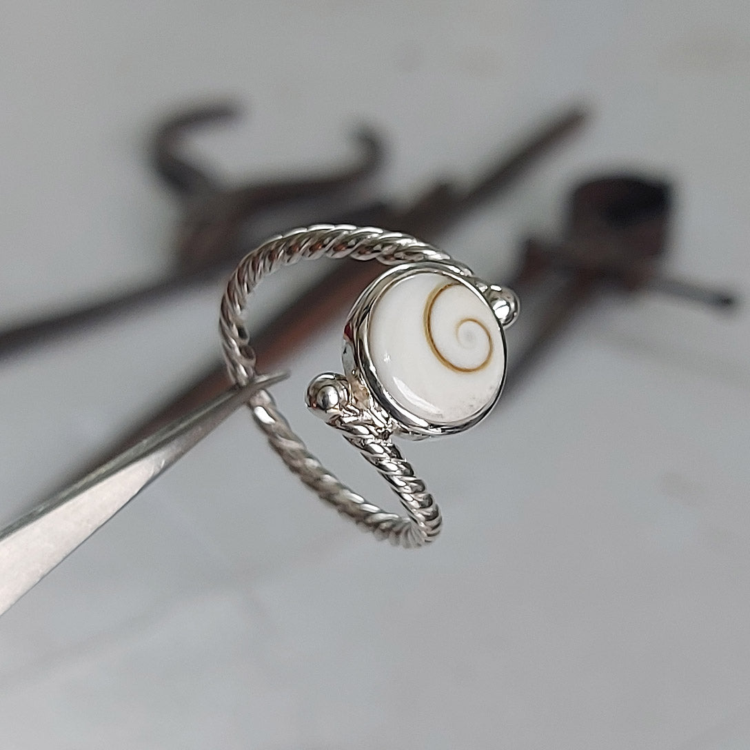 A silver ring with a twist design featuring a Shiva's eye pendant in the center, displayed on a tool-like surface.