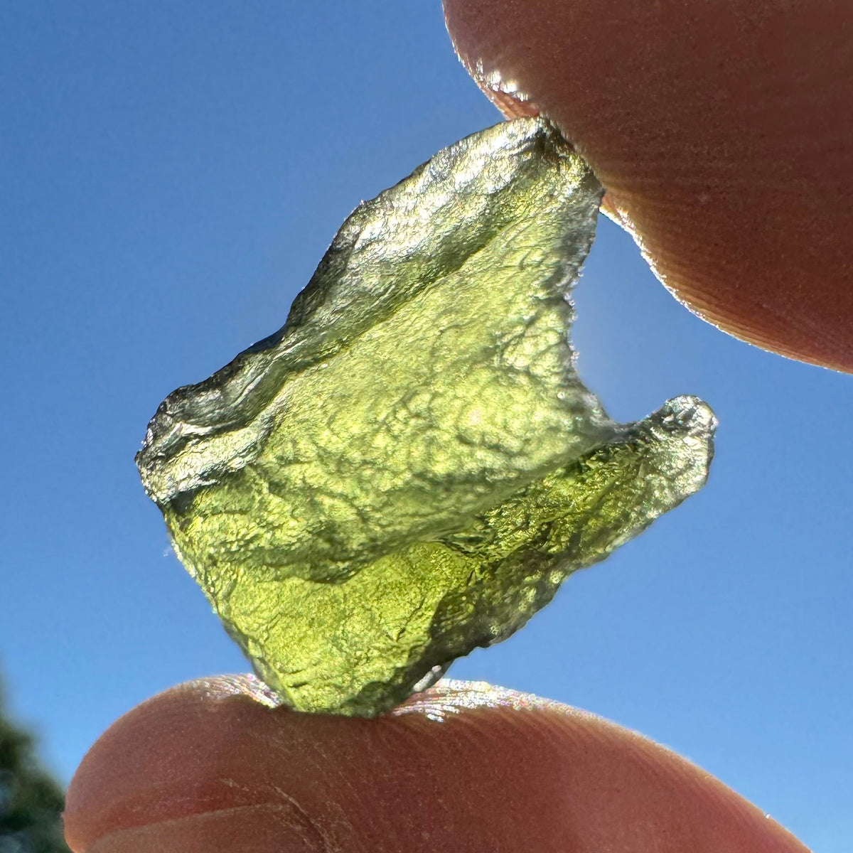 A translucent olive-green glass fragment, likely molten glass, held between two human fingers against a bright blue sky with blurred outdoor background.