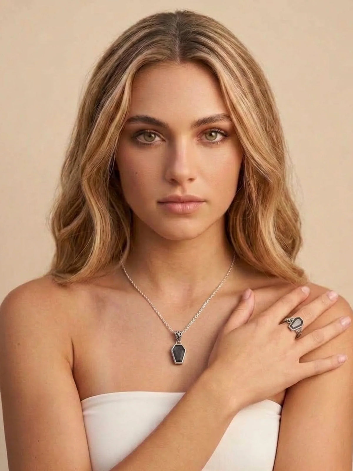 A woman with blonde hair wearing a silver chain necklace with a dark gray geometric pendant and a matching silver ring, posing against a solid beige background.