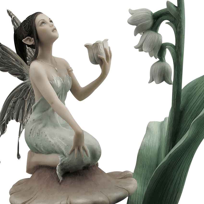 The Lily of the Valley Statue By Rachel Anderson captures a charming scene in which a fairy waits patiently for a drop of dew to fall from one bell-shaped flower. Sitting atop a brown toadstool, the fairy wears a light green tunic with a chain of pearls and a teardrop pendant hanging from her neck. 