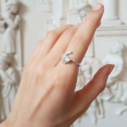 A hand holding a sterling silver ring with a crescent moon moonstone design, against an ornate background.